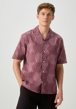 Ung mand iført en kortærmet skjorte med knapper og et mønster af lilla bølgende linjer, stående foran en ensartet lys baggrund.