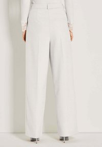 Vrouw in hoge taille, wijde witte broeken, gecombineerd met een geribbelde witte top met kant aan de manchetten, staande tegen een effen muur.