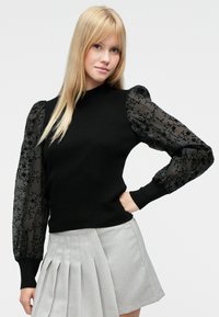 Schwarzer, gerippter Pullover mit durchsichtigen, schwarz bestickten Puffärmeln mit Blumenmuster. Kombiniert mit einem plissierten, hellgrauen Rock.