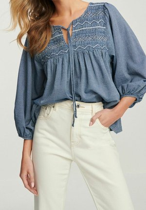 Femme portant un chemisier bleu smocké à manches longues bouffantes et un pantalon taille haute blanc cassé, main dans la poche.