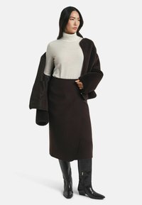 Brauner Wollwickelrock kombiniert mit einem hellen Rollkragenpullover und einer dunklen, strukturierten Außenjacke. Glänzende schwarze kniehohe Stiefel mit Krokodilprägung.