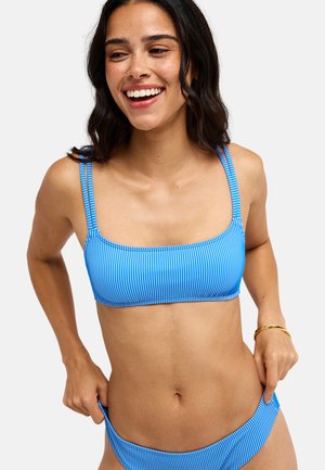 Femme souriante aux cheveux foncés portant un bikini rayé bleu et blanc, ajustant le bas avec les deux mains.