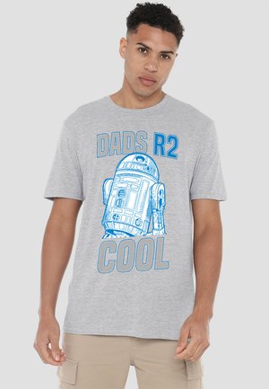 Young man wearing a light gray T-shirt with blue R2-D2 robot illustration and the text "DADS R2 COOL" standing against plain background.