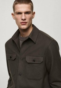 Dark green button-up jacket made of smooth fabric, featuring two chest pockets with black buttons and a pointed collar.
