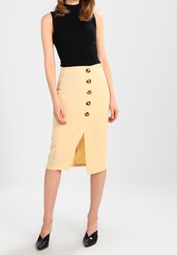 Žlutá sukně na tužku s předním rozparkem, zdobená pěti kulatými hnědými knoflíky uprostřed, kombinovaná s přiléhavým černým topem bez rukávů.