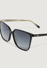 Occhiali da sole con montatura nera a quadrato, lenti grigie sfumate e bracci con dettagli dorati che presentano un piccolo logo rosso, bianco e blu.