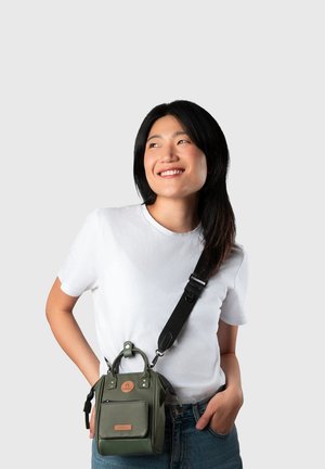 Junge Frau lächelt, trägt ein weißes T-Shirt und blaue Jeans, und hat eine kleine grüne Umhängetasche mit verstellbarem schwarzem Riemen dabei.