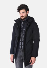 Parka nera impermeabile con cappuccio, indossata sopra un piumino scuro. Presenta una chiusura con zip e diverse tasche, mostrata su un modello.
