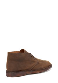 Botín chukka de cuero marrón con textura suave, punta redonda, dos ojales y suela marrón en contraste. Presenta un diseño minimalista.