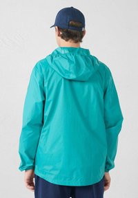 Tijlgroene lichtgewicht jas met capuchon, elastische manchetten, een ontspannen pasvorm en een trekkoord aan de capuchon, gedragen over marineblauwe broek. Gladde, waterbestendige stof.