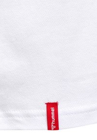 Bílá texturovaná látka s červenou štítkem, na kterém je vyšitý název značky "hummel" na dolním okraji.