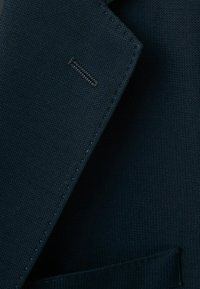 Dark navy blue textured fabric close-up showing the notch lapel and stitched buttonhole of a suit jacket.