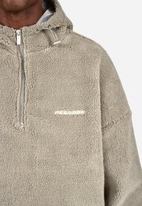 Világos bézs színű bolyhos pulóver fél cipzárral és kapucnival, amely texturált felülettel és diszkrét márkázással rendelkezik a mellkasán.