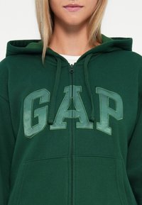 Zelena dukserica sa patentnim zatvaračem i mekim materijalom, s velikim natpisom "GAP" u svjetlijoj zelenoj boji na prednjem dijelu. Ima kapuljaču i prednje džepove.