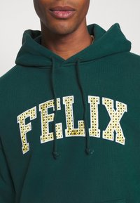 Felpa con cappuccio verde scuro caratterizzata da un logo "FELIX" prominente in bianco e giallo con dettagli decorativi. Coulisse sul cappuccio. Materiale in cotone.