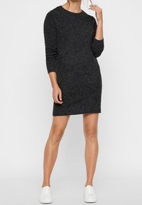 Kvinna iklädd en långärmad, mörkgrå stickad klänning och vita sneakers, står mot en ljus, enfärgad bakgrund.