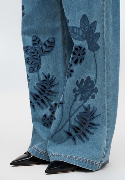 Τζιν παντελόνια με σκούρο μπλε κεντήματα με λουλούδια και σχέδια με κοψίματα, με μυτερά μαύρα τακούνια ορατά στο κάτω μέρος.