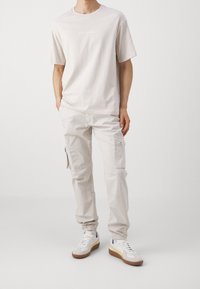 Mężczyzna ubrany w jasnobeżowe spodnie cargo, dopasowaną oversize'ową koszulkę oraz białe sneakersy z żółtym paskiem i gumową podeszwą, stoi z rękami w kieszeniach.