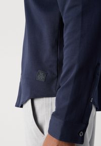 Maglietta a maniche lunghe blu navy con tessuto testurizzato, caratterizzata da un evidente logo applicato, polsini con bottoni e aperture laterali.