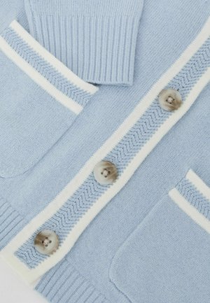 Lichtblauwe cardigan van gebreide stof, met witte bies, visgraatmotief en grote bruine knopen. Voorzien van twee zakken aan de voorkant.