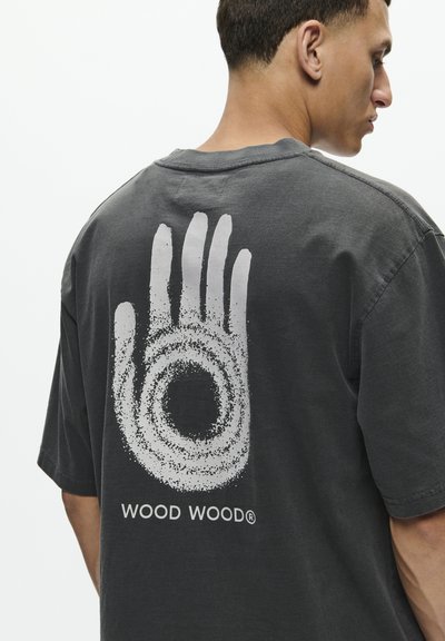 Pelēks kokvilnas T-krekls ar lielu baltu rokas grafiku ar apaļu dizainu un tekstu "WOOD WOOD" apakšā aizmugurē.