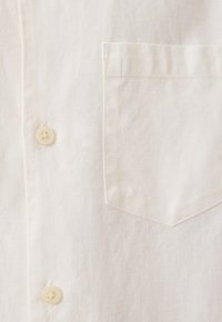 Lyse beige linned skjorte med en struktureret finish, der har en knaplukning foran og en venstre brystlomme. Knapperne er runde og cremefarvede.