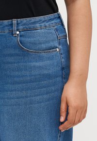 Μπλε τζιν φούστα με ψηλή μέση, διαθέτει ασημί μεταλλικά στοιχεία, μπροστινές τσέπες και λεί�α υφή. Χέρι που ξεκουράζεται στο ύφασμα.