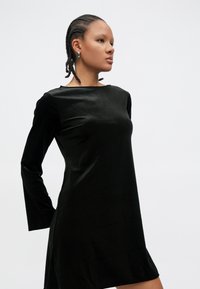 Vestido de veludo preto com mangas compridas e decote canoa, apresentando um corte reto e textura suave, que termina acima dos joelhos.