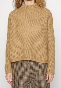 Maglione lavorato a maglia color beige con collo alto e motivo testurizzato. Vestibilità ampia e polsini a costine. Indossato sopra pantaloni a quadri marroni.