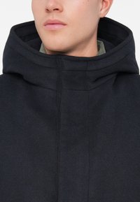 Osoba v tmavom námorníckom fleecovom kabáte s vysokým golierom a kapucňou, ktorá čiastočne zakrýva svetlozelený golier košele.