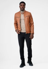 Brun læderjakke med opretstående krave, sidelommer og brystlommer med lynlås. Båret over en beige turtleneck, parret med sorte slim-fit bukser og støvler.