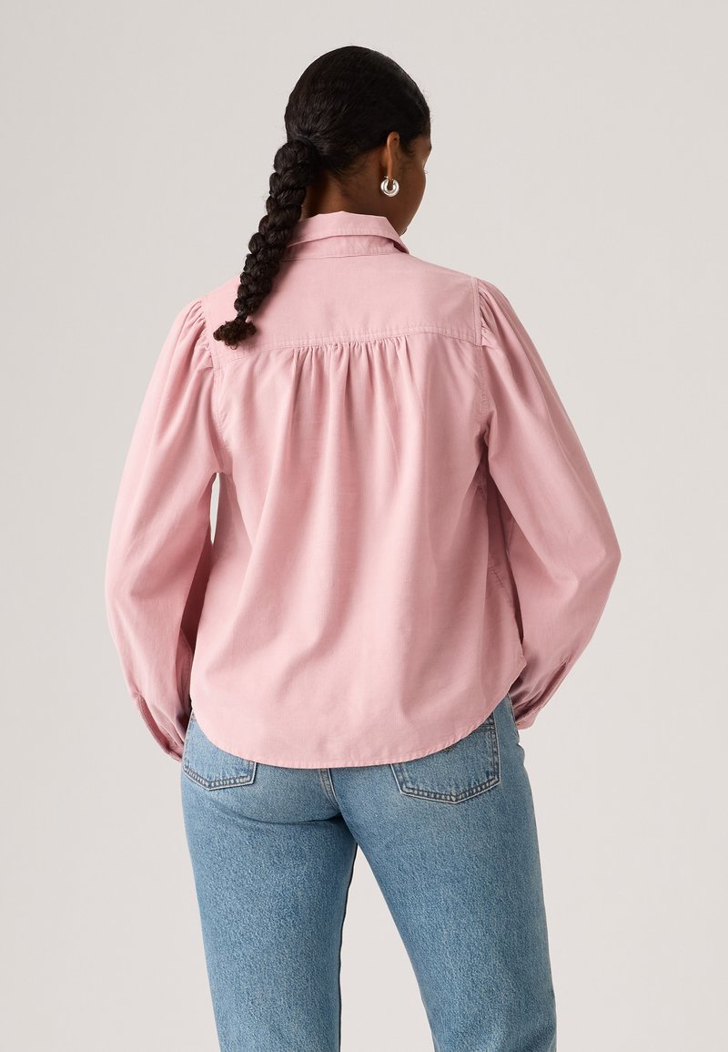 Chemise rose clair à manches longues avec col boutonné et détail froncé au dos. Associée à un jean en denim bleu, avec une légère courbure à l'ourlet.