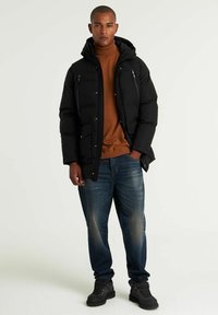 Svart vadderad jacka med huva, blixtlås och tryckknappar, två framfickor. Kombinerad med en brun polotröja och blå jeans, avslutad med svarta stövlar.