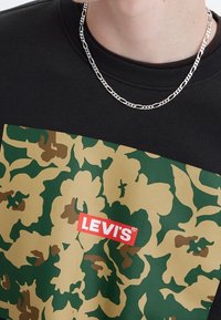 Felpa nera con un motivo floreale verde e beige, con un’etichetta rossa "LEVI'S" in evidenza. Una collana a catena aggiunge un tocco metallico.