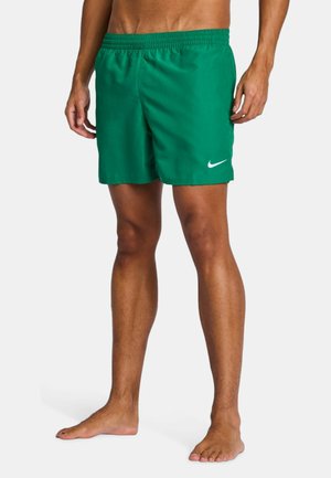 Modello maschile che indossa pantaloncini da bagno verdi Nike con logo bianco, in piedi a piedi nudi su sfondo semplice.