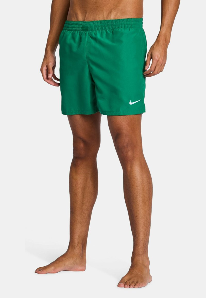 Modèle masculin portant un short de bain Nike vert avec logo blanc, debout pieds nus sur un fond uni.