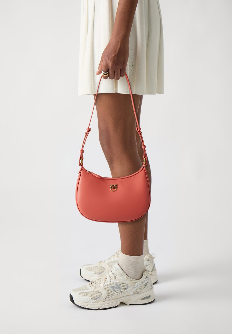 Mala coral com uma textura suave e forma curva, apresentando acessórios dourados e uma alça ajustável, segurada numa mão perto de um vestido branco.