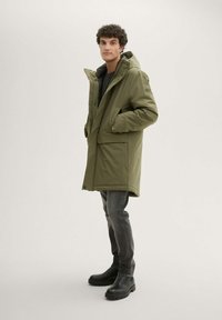 Parka isolata verde oliva con cappuccio, dotata di chiusura con zip, due tasche frontali e una vestibilità comoda. Indossata sopra un maglione scuro e jeans grigi.