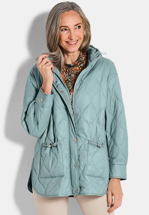 Lächelnde Frau mittleren Alters mit grauen Haaren, die eine hellblaue gesteppte Jacke über einem gemusterten Oberteil trägt, vor einem schlichten hellen Hintergrund stehend.