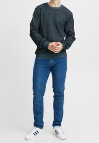 Persona con felpa grigia scura, jeans blu e sneakers bianche con strisce nere, in piedi su uno sfondo chiaro semplice.
