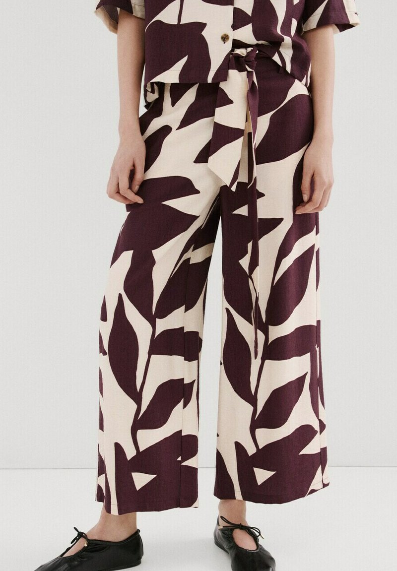 Pantaloni cu croială largă, cu model mare de frunze în nuanțe de violet închis și crem, cu centură legată în talie, asortați cu top și balerini negri.