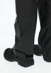 Pantaloni neri impermeabili con texture liscia, dotati di una fascia in velcro alla caviglia, abbinati a scarpe nere robuste con suole scolpite.