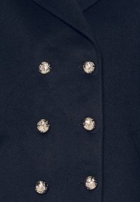 Manteau bleu marine croisé en laine, avec des boutons métalliques argentés ornés de motifs en relief et une texture lisse.