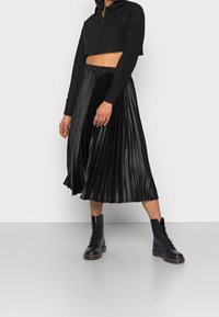 Black cropped button-up shirt paired with a pleated black midi skirt made of shiny material, and black lace-up combat boots.