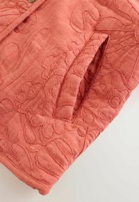 Közelkép korallszínű anyagról, virágmintás hímzéssel és hasítékos zseb részlettel.