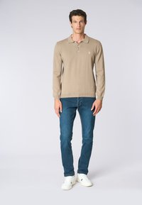 Polo beige a maniche lunghe con vestibilità rilassata, dotato di colletto e patta con tre bottoni. Indossato con jeans blu scuro e sneakers bianche.