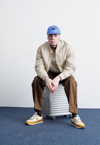 Beige Bomberjacke über einem cremefarbenen Hemd, braunen Hosen und mehrfarbigen Sneakern. Blaue Mütze und sitzt auf einem silbernen Metallkoffer.