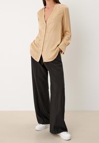 Chemisier en satin beige à manches longues avec un décolleté en V et des fentes latérales, associé avec un pantalon large noir et des baskets blanches.