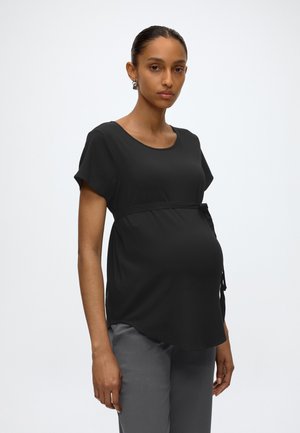 Gravid kvinde iført en sort top med korte ærmer og taljebånd samt grå bukser mod en ensfarvet lys baggrund.