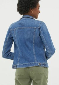 Woman wearing a blue denim jacket and green pants, turned slightly to the right, smiling with hands near her waist.